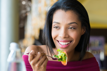 Closeup shot of young woman eating fresh salad at restaurant. Healthy african girl eating salad and looking away. Smiling young woman holding a forkful of salad. Health and diet concept. Woman ina a lunch break.の写真素材