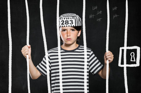 Closeup of sad bad boy wearing jail suit and kept behind bars. Portrait of little prank in jail. Sad male kid wearing striped clothes.の写真素材