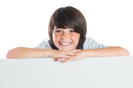 Portrait of smiling little boy leaning on placard isolated on white background. Young boy leans over poster. Happy cute kid leaning on blank white poster. Closeup of smiling boy leaning on empty grey placard and looking at camera.の写真素材