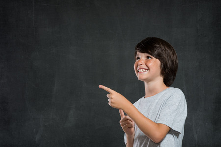 Closeup of little boy pointing finger isolated on blackboard. Happy smiling boy looking up and pointing. Portrait of cheerful boy showing on blackboard, copy space for your text or product.の写真素材
