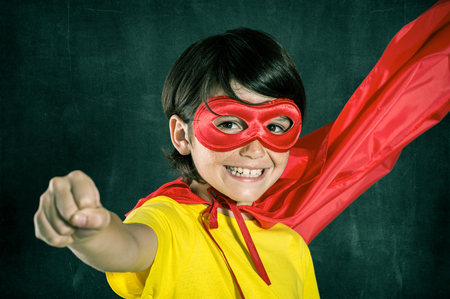 Closeup of little boy in superhero costume posing isolated on blackboard. Smiling kid looking at camera with red mask. Cheerful cute boy wearing superhero costume with red cape flying with closed fist.の写真素材