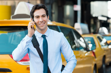 Young businessman talking on smartphone. Happy young casual professional businessman using cell phone outdside, in a city centre. Portrait of a handsome young man talking on the phone with yellow taxi in background.の写真素材