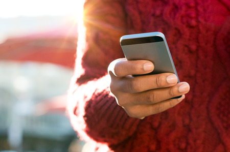 Close up of a young woman's hand typing text message on her smartphone. African young woman is typing on touch screen mobile phone. Close up of female hand texting a message phone outdoor.の写真素材