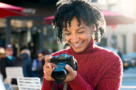 Portrait of african tourist with digital camera at cafe. Joyful woman looking at pictures on digital camera sitting outside the cafetria. American woman taking picture photo with camera ih her holiday.の写真素材