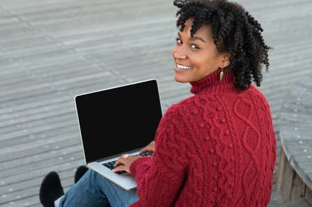 Young african woman sitting on wooden stairs outdoor and working on laptop. Happy casual woman sitting on wooden floors with laptop and looking at camera. Smiling adult student studying on pc.