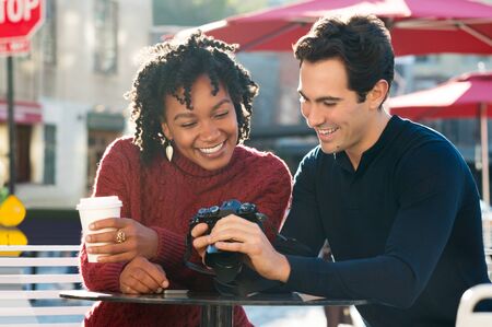 Happy young tourist couple looking at pictures in their camera in a cafe. A smiling couple looking at their photos on the screen of their camera in the cafe outdoor. Joyful multi ethnic couple sitting at cafeteria using a camera with coffee paper cup in hの写真素材