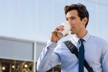 Successful young businessman drinking a take away coffee outside office. Businessman drinking coffee and thinking about his future. Happy ambitious man drinking an hot coffee with a paper cup in front of the modern building.の写真素材