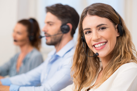 Smiling female call centre operator doing her job with a headset while looking at the camera. Portrait of happy woman in a call center smiling and working. Portrait of happy smiling female customer support phone operator at workplace.