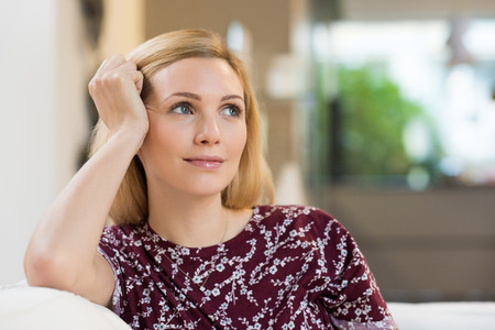Young blond woman leaning her head against her hand while sitting on a white sofa and looking away.の写真素材