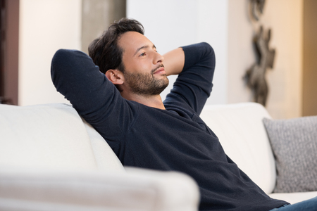 Handsome young man with hands behind head sitting on couch in living room.