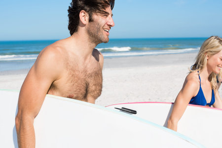 Joyful couple with surfboards walking towards the sea. Young couple running on beach with surfboards under their arms. Smiling young active surfers couple with surfboards on the beach.の写真素材
