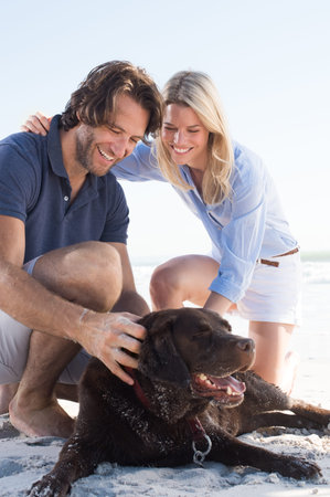 Cheerful couple stroking labrador retriever at beach. Happy smiling couple playing with their dog at...