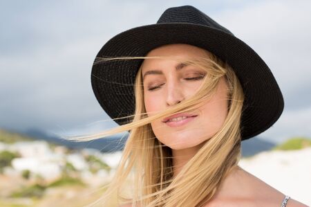 Close up face of a beautiful woman wearing black strawhat on a bright sunny day with her eyes closed. Young blonde woman with closed eyes enjoying sunny day. Woman relaxing at beach.の写真素材