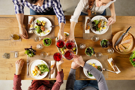 Top view of group of friends raising a toast post lunch. High angle view of happy men and women celebrating at home with red wine. Close up shot of friends toasting glasses of red wine in a party.