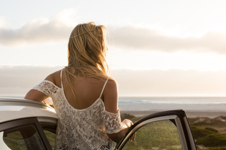Young traveller woman enjoying sunset standing outside car. Serene woman leaning on car during sunset. Young woman on summer travel to the coast.の写真素材