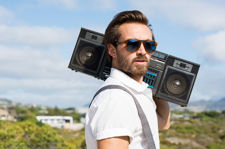 Portrait of a handsome young man holding radio near ears. Close up of hipster guy listening to music on a summer day. Man wearing sunglasses and holding on his shoulder a vintage radio.の写真素材