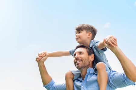 Father giving son ride on back in park. Portrait of happy father giving son piggyback ride on his shoulders and looking up. Cute boy with dad playing outdoor.