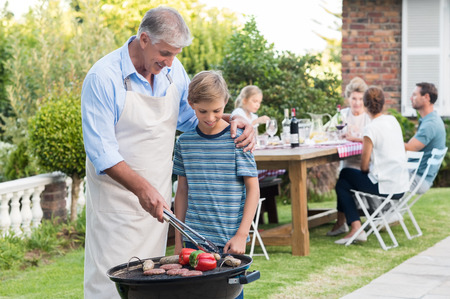 Grandson helping grandfather in barbeque. Happy grandpa teaching grandson how to cook food. Grandson enjoying watching grandfather cooking barbeque.の写真素材