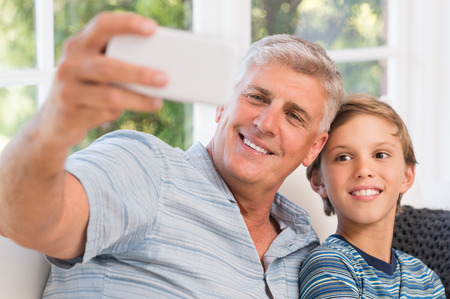 Smiling grandfather with grandson taking selfie with mobile phone. Grandchild taking selfie with smiling grandpa. Portrait of grandfather with grandson sitting on sofa at home.の写真素材