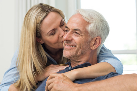 Senior woman embracing her husband from behind. Close up face of mature woman kissing man on cheek in living room. Portrait of a loving wife kissing senior man.の写真素材