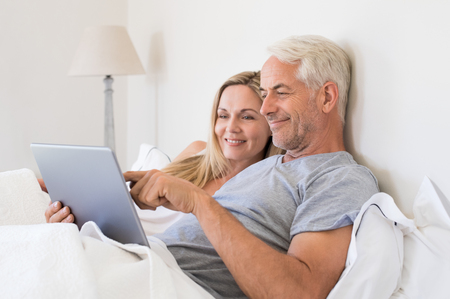 Senior couple using digital tablet while lying in bed at home. Happy senior couple surfing on the tablet in bedroom. Retired man and smiling woman planning on laptop their summer vacation.の写真素材