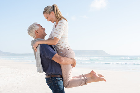 Cute senior couple hugging on the beach on a sunny day. Happy couple having fun together at the beach. Senior man carrrying her wife at beach and looking at each other.