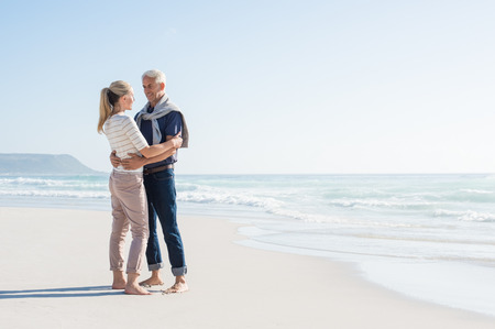 Cheerful mature couple embracing on the beach with copy space on right. Happy senior couple looking at each other at seaside. Romantic retired couple on summer vacation at sea.の写真素材