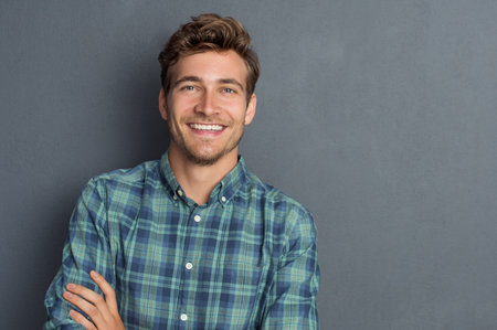 Young handsome man leaning against grey wall with arms crossed. Cheerful man laughing and looking at...