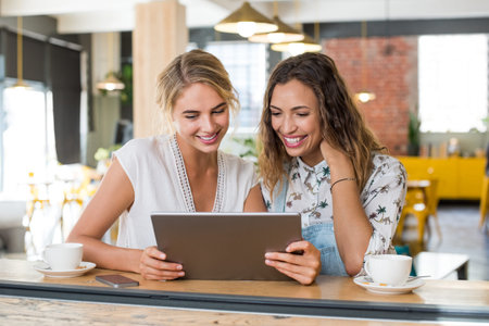 Two happy smiling young women in a cafeteria using digital tablet. Happy girls friends sitting at coffee shop looking at tablet. Happy multiethnic women drinking coffee while using tablet.の写真素材