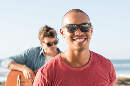 Portrait of a smiling man wearing sunglasses in front of man playing guitar. African guy looking at camera during sunset. Portait of carefree black man in casual smiling.の写真素材