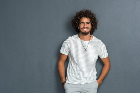 Portrait of happy young man with curly hair standing against grey background. African man with hands in pocket leaning against grey wall. Multi ethnic young man in casual looking at camera with copy space.の写真素材