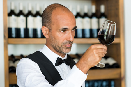Professional sommelier examining wine in a glass. Bartender examining red wine in glass at shop. Multiethnic sommelier examining glass with red wine.の写真素材