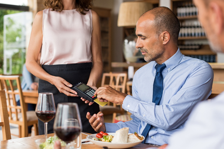 Waiter holding credit card swipe machine while customer typing code. Mature businessman making payment in cafe through credit card. Customer paying bill of lunch with debit card.の写真素材