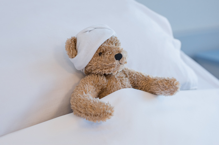 Injured teddy bear sleeping on hospital bed. Brown plush toy with a headache resting on bed. Sick brown bear with a bandage on his head lying in bed hospitalized at medical clinic.の写真素材