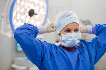 Young doctor during preparation for operation. Assistant of surgeon wearing surgical mask in operation room at the hospital. Female surgeon attaching his mask in an operating theater.の写真素材
