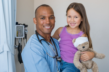 Happy male nurse carrying cute girl in hospital room. Doctor with stethoscope and little patient looking at camera in a medical clinic. African man with young child patient smiling after medical exam.の写真素材