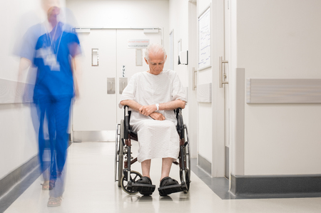 Sad old man sitting on wheelchair in hospital corridor. Depressed senior patient in hospital hallway while nurse passing by. Blur motion of nurse passing beside a elderly patient with alzheimer.の写真素材