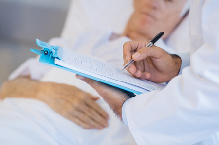 Close up on doctor hand writing on clipboard. Senior doctor hand filling on medical presciption at hospital. Closeup of hand of a doctor holding medical report with ill patinet lying on bed in background.の写真素材