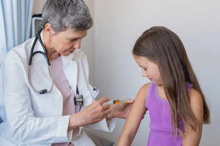 Senior doctor vaccinating small girl at hospital. Pediatrician giving child an injection in arm for flu vaccine. Little patient receiving injection in exam room.の写真素材