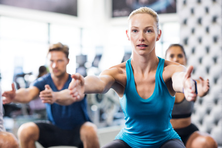 Young woman exercising in gym with people in background. Fit woman exercising with stretched hands and squats in modern gym. Fitness class exercising in gym.の写真素材