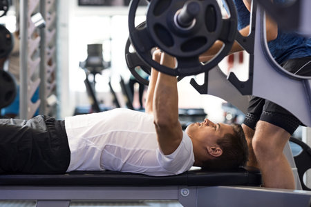 Determined young man lifting barbell. Latin man doing barbell bench press in gym with personal...
