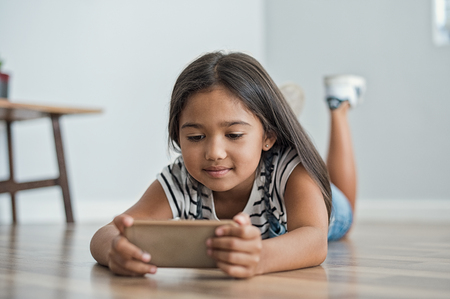 Little asian girl lying on floor using mobile phone to play. Multiethnic female child watching cartoon on smartphone at home. Childhood and moder technology concept.の写真素材