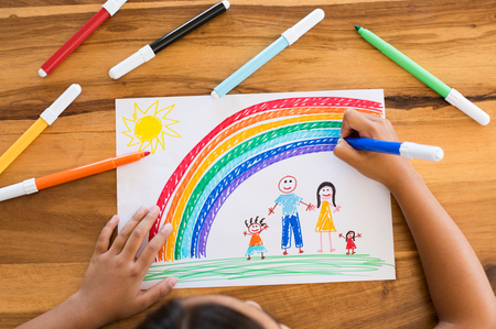 Top view of little girls hands painting happy family with marker on white paper. Drawing sheet with artwork by daughter on table. Little girl using marker to make family painting with rainbow.の写真素材