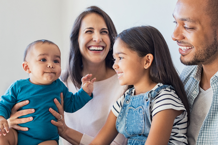 Happy daughter sitting on father legs and looking his little brother. Excited infant playing with elder sister. Portrait of smiling multiethnic family sit on sfoa and enjoying at home.の写真素材