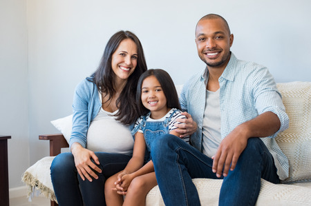 Happy multiethnic couple with their daughter sitting couch at home. Smiling family with one child expecting another baby. Pregnant mother sitting with daughter and black husband on couch.の写真素材