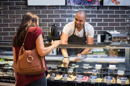 African waiter serving fresh food to young woman. Happy smiling guy preparing take away salad for woman customer. Black man with apron taking pasta salad in spoon and serving to customer.の写真素材