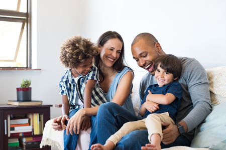 Happy multiethnic family sitting on sofa laughing together. Cheerful parents playing with their sons at home. Black father tickles his little boy while the mother and the brother smile.の写真素材