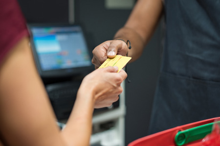 Close up of customers hand giving credit card to cashier at checkout. Woman using credit card to pay at supermarket. Woman paying with debit card at cash register.の写真素材