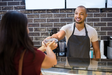 Young waiter giving parcel to customer at counter in cafeteria. Young waiter at cafe giving take away salad to woman customer. Happy african man selling food to woman.の写真素材