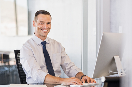Portrait of confident manager sitting at desk and looking at camera. Portrait of smiling business man working at computer. Successful formal man in his new modern office.の写真素材